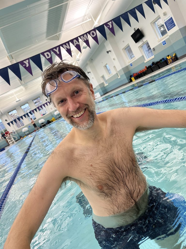 A tilted photo of Ken standing in a pool with flags spelling DYNAMO above and behind him