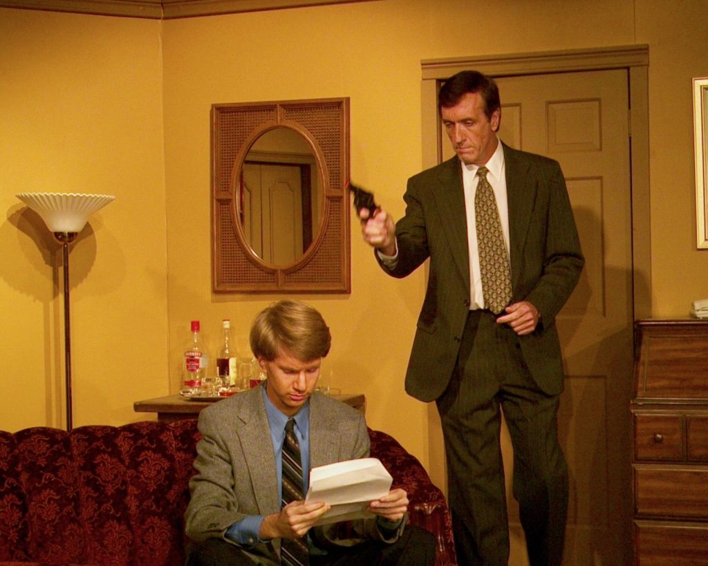 Ken sitting on a couch, reading a letter, as someone behind him points a gun at his head