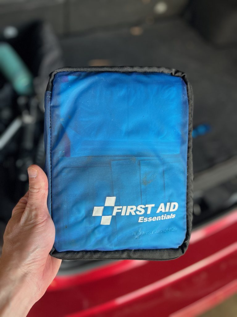 A hand holding a small blue pouch labeled "First Aid Essentials"